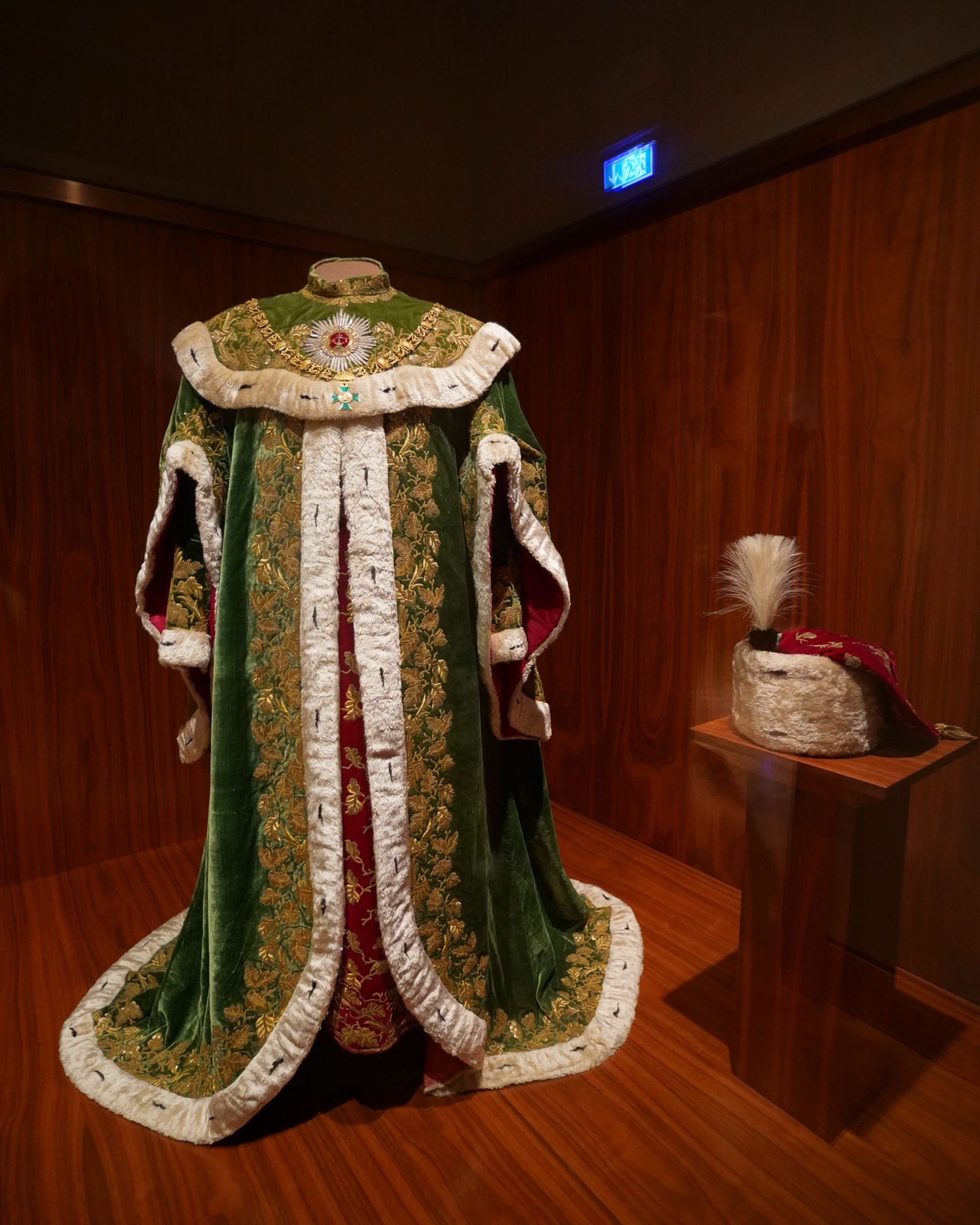 A full-length robe of office, green and gold and lined with white ermine; a plumed hat is on a stand nearby