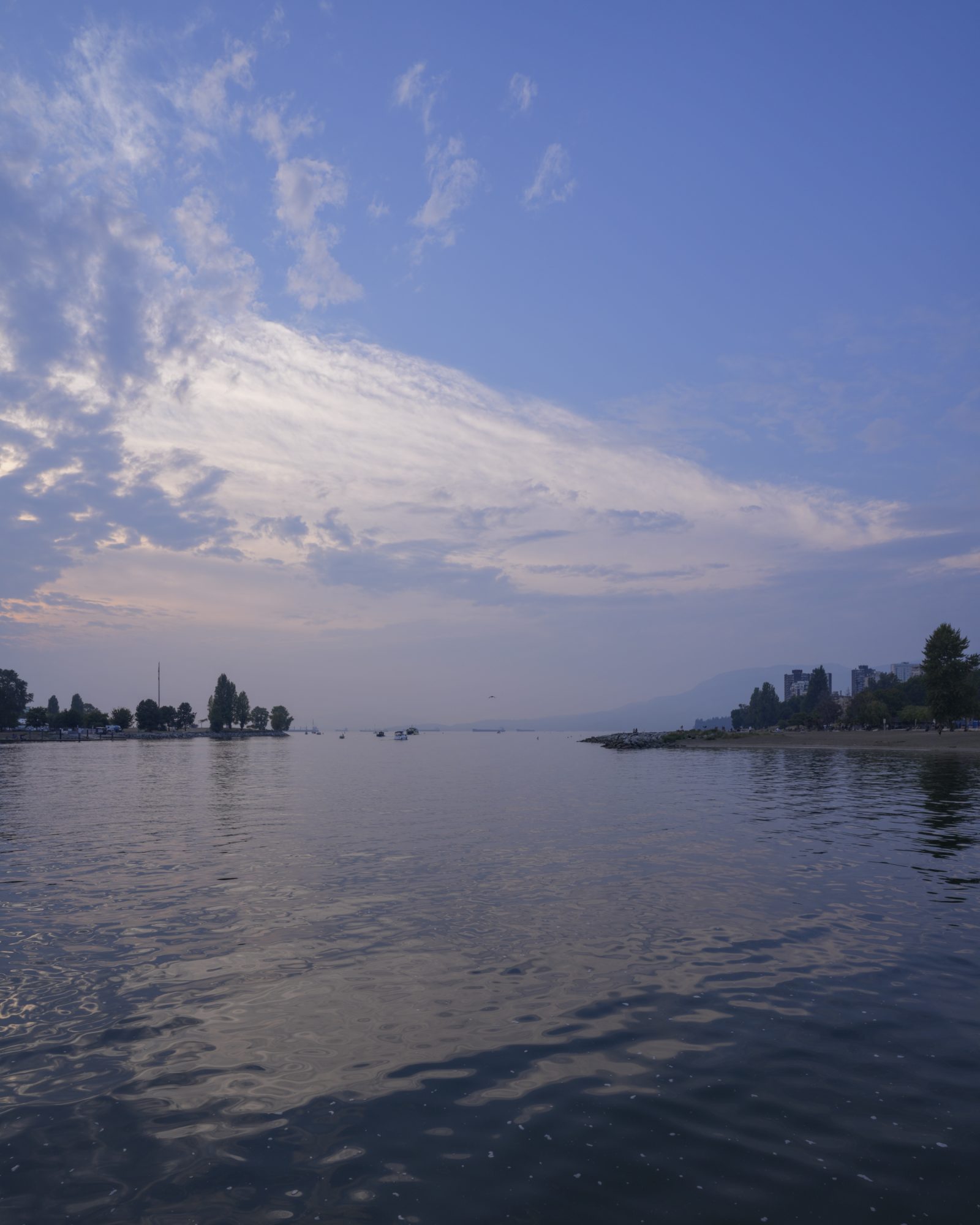 View from Sunset Beach, over English Bay. The sky is mostly blue with some small clouds, and the water is still. There is greyish haze on the horizon, partially hiding the mountains