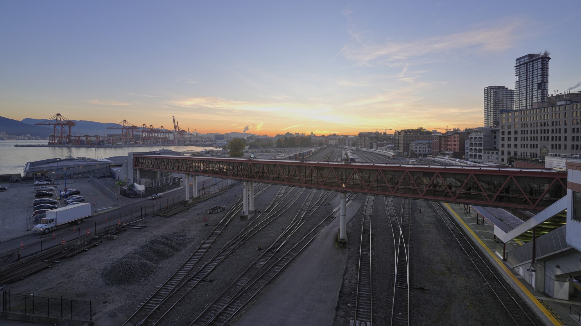 View east from Canada Place, at sunrise. We see the walkway to the SeaBus terminal, the train tracks, big cranes in the distance