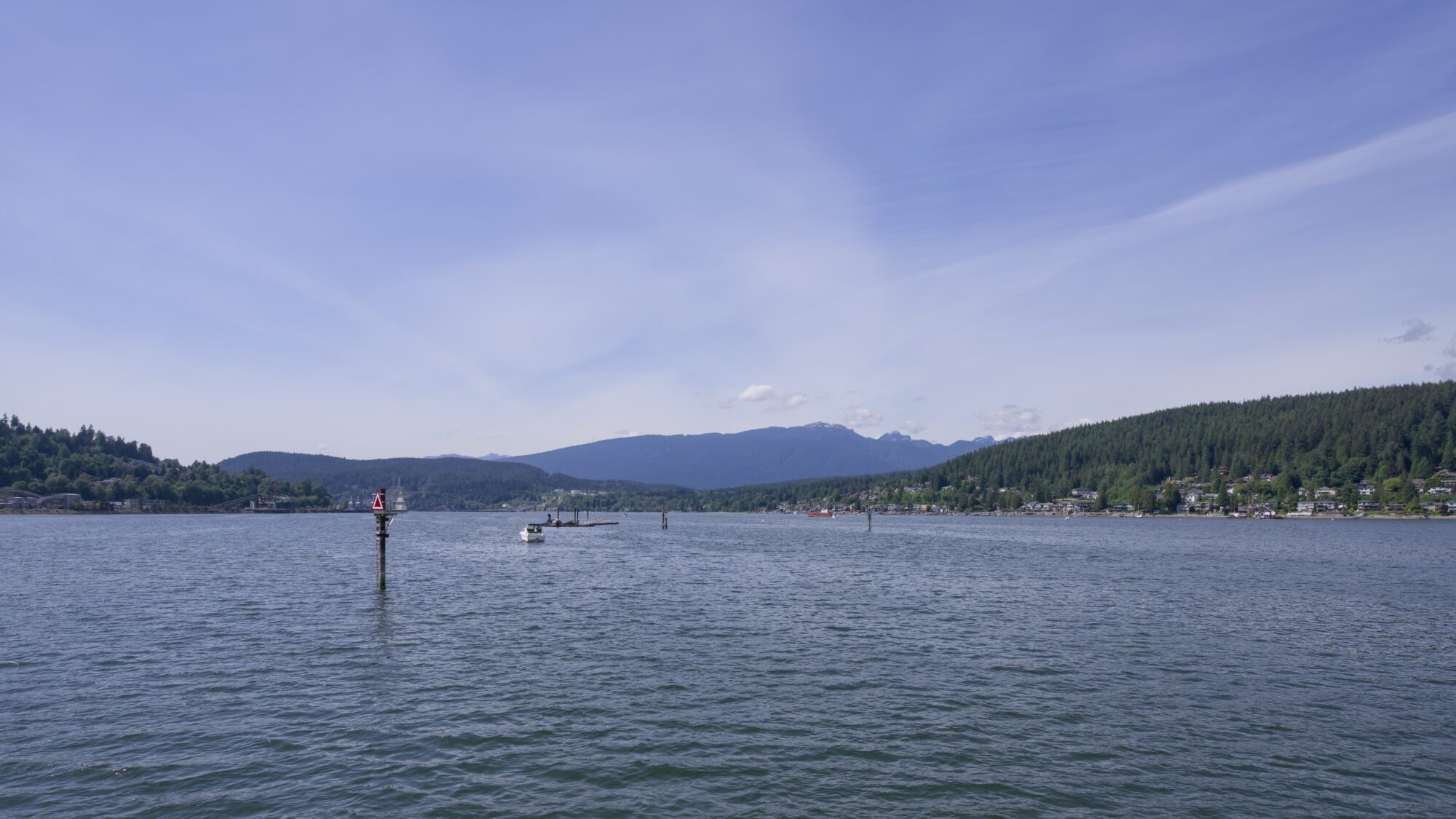 View from the Rocky Point Park pier: some pilings, and water off in the distance between low rolling mountains under a blue sky with wispy clouds